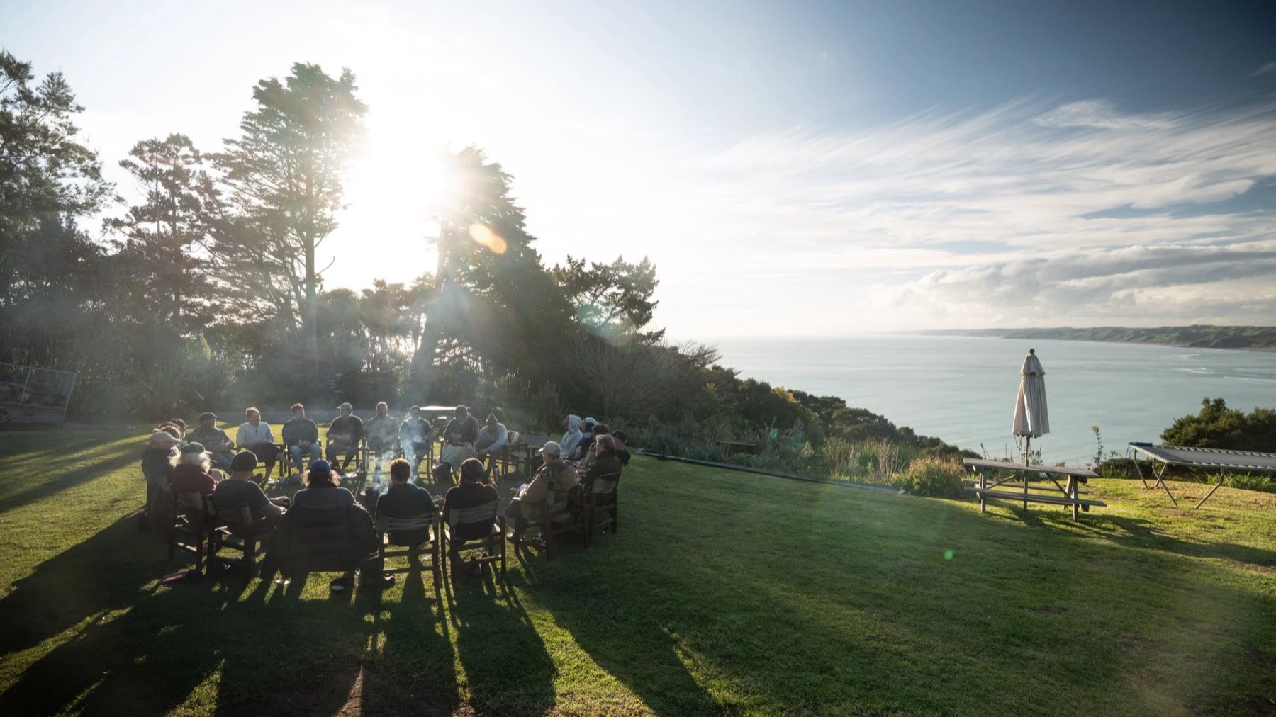 A circle of men around a fire at golden hour, overlooking the sea